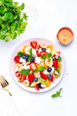Summer fruit and berry salad with fresh strawberries, blueberries, banana, cottage cheese and mint on white table background, top view