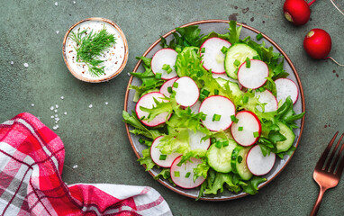 Healthy crispy salad with radishes, cucumbers, lettuce and chives with yogurt dressing, green stone table background, top view