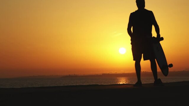 Silhouette of a man near the ocean with a longboard at bright orange sunset