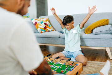 happy redhead father and son smiling at camera while playing table football together at home