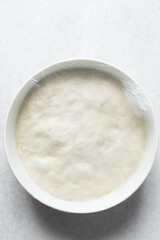 Plastic wrap covered bread dough that has doubled in size , dough that has fermented, proofed dough in a white bowl , process of making focaccia