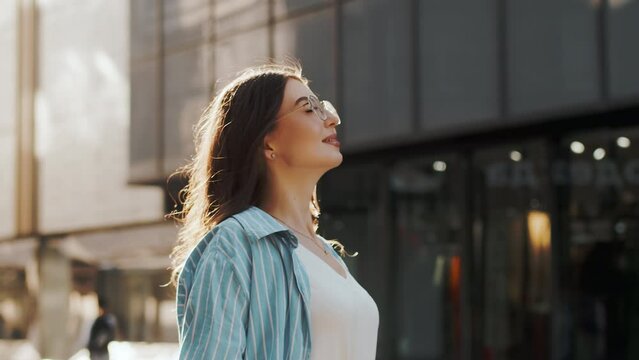 Young beautiful woman in casual clothes takes a deep breath looking at the sun and enjoying summer on the city street. Concept of an urban carefree life