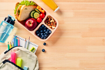 School lunch box with sandwich, fruits, vegetables, bottle of water, and kids backpack with school supplies on wooden table. Flat lay, top view.