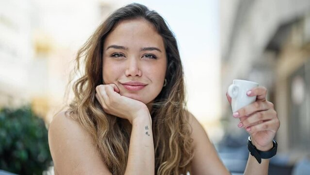 Young beautiful hispanic woman sitting on table drinking coffee smiling at coffee shop terrace