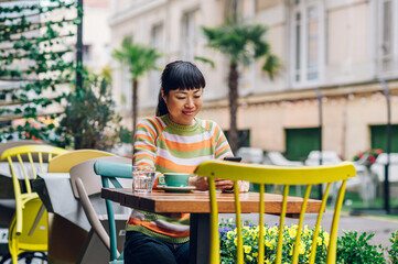 Joyful asian woman sitting in an outdoor restaurant and looking at her phone.