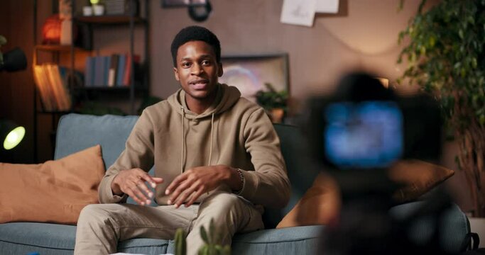 Motivational speaker African American man sits in living room addressing camera with confidence passion holding notepad pen shares inspiring messages of personal growth and resilience.