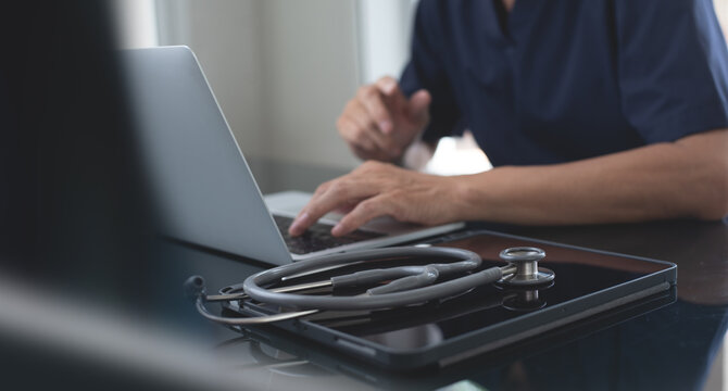 Close up of male doctor working and typing on laptop computer at doctor's office with stethoscope and digital tablet on table, doctor staff online meeting via laptop