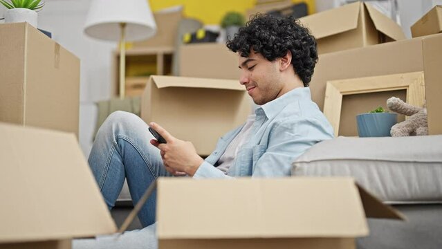 Young latin man using smartphone sitting on floor smiling at new home