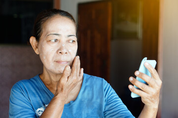 Asian senior woman apply  anti aging facial cream on her face, holds and look at small mirror....
