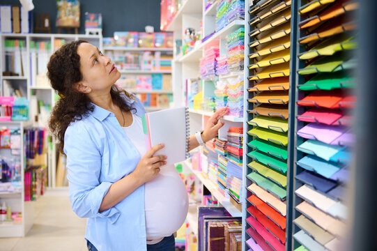 Multi-ethnic pregnant woman, inspired artist painter standing by shelf with color paper sheets in school stationery shop