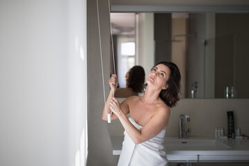 Middle-aged woman opening white roller blinds on window in a modern apartement