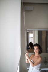 Middle-aged woman opening white roller blinds on window in a modern apartement