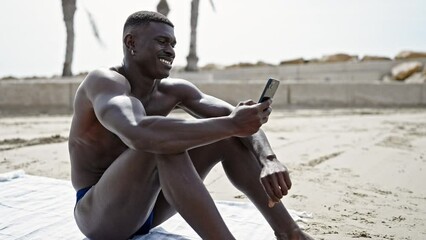 African american man tourist sitting on towel shirtless using smartphone at the beach