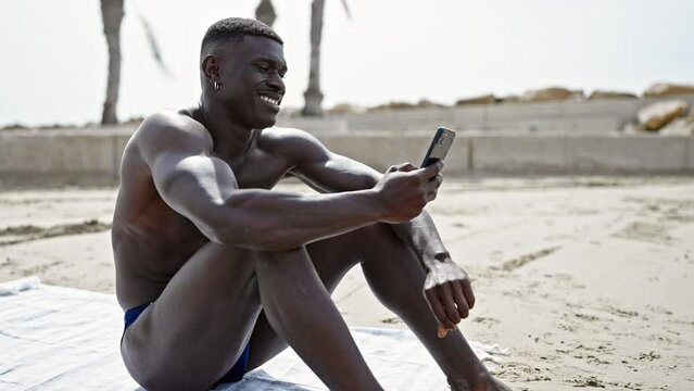 African american man tourist sitting on towel shirtless using smartphone at the beach