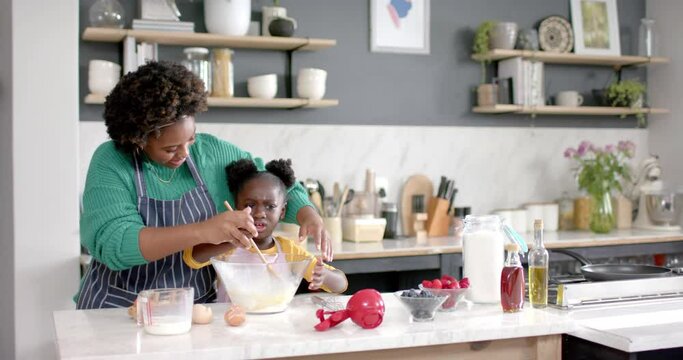 Happy african american mother and daughter preparing dough in bowl in kitchen, slow motion