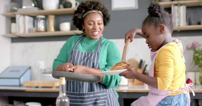 Happy african american mother and daughter frying pancakes in kitchen, slow motion