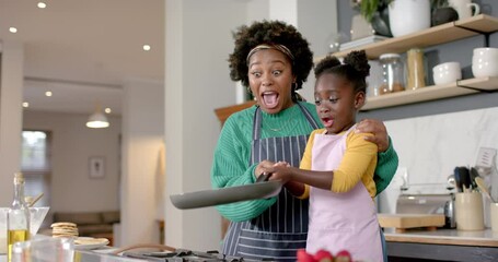 Happy african american mother and daughter frying pancakes in kitchen, slow motion