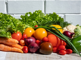 A pile of fresh, healthy and colorful vegetables in bright sunlight with white brick wall in background. Tomatoes, onions, carrots, peppers, cauliflower, salad, dill and greens