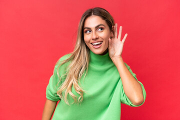 Young Uruguayan woman isolated on red background listening to something by putting hand on the ear