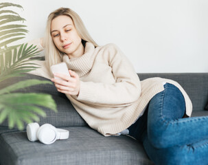 Happy young woman relaxing on comfortable couch, holding smartphone in hands.
