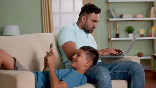 Indian father working on laptop while son using mobile phone by sleeping on lap on sofa at home - concept of Digital connection, technology addiction and cyberspace.