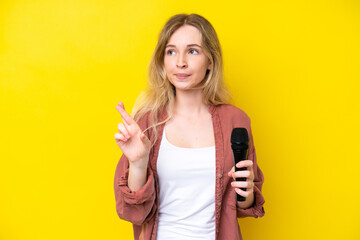 Young singer caucasian woman picking up a microphone isolated on yellow background with fingers crossing and wishing the best