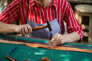 Closeup and crop hands of young female leather maker using a small hummer with leather punching...