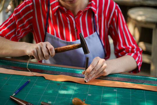 Closeup and crop hands of young female leather maker using a small hummer with leather punching steel to make a leather belt for customers. - Powered by Adobe
