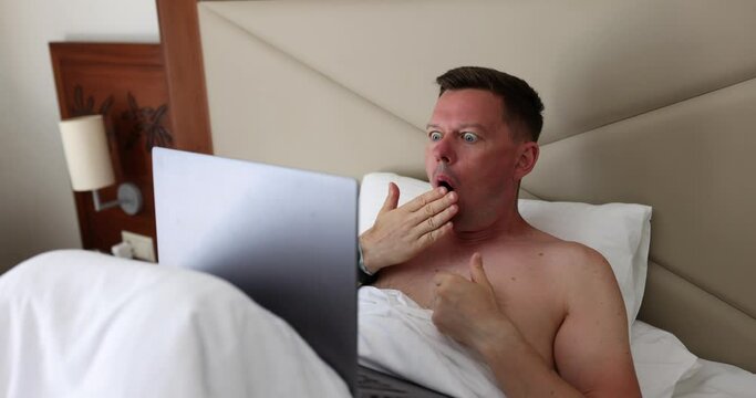 Man using laptop lies in bed, covering mouth with hand shocked and afraid of making mistake. Emotions of surprise and surprise
