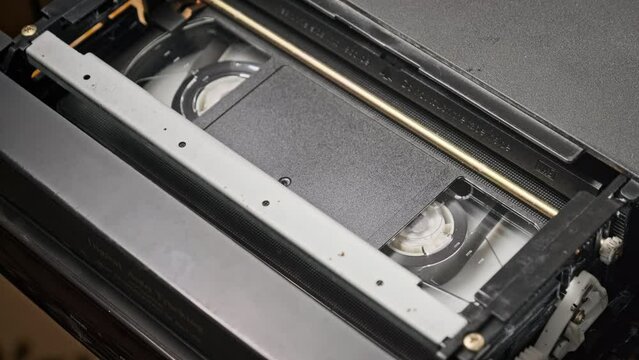 VHS cassette is played inside a VCR tape recorder, top view. Videotape reels rotate. Videocassette with empty blank tag is starting playback. Old video recorder inside close-up. Playing vintage movie