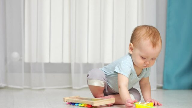 Baby girl plays around with xylophone toy on floor in house. Small child in ordinary clothes learning to play music using colorful toys