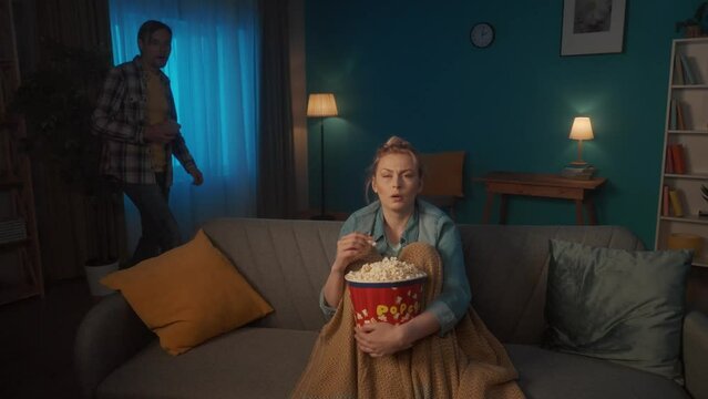Shocked woman watching a scary movie on TV at home and eating popcorn. A man with a phone in his hands comes up to a woman from behind and scares her. A man hugs a woman and kisses her soothingly.