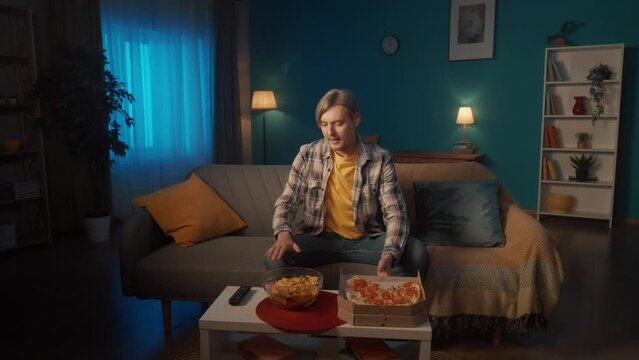 A man is preparing to watch a movie. Brings a plate of chips and boxes of pizza. The man sits down on the couch, turns on the movie with the remote control. A man is enjoying a comedy movie at home.