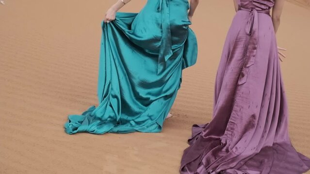 Two young women in long dresses developing in the wind run barefoot through the sand dunes of the desert