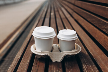 Takeaway paper coffee cups with lids in cardboard holder on wooden bench outdoors. White paper cups of coffee mockup