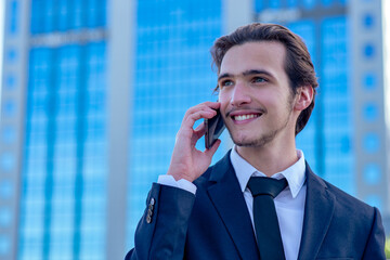 Smiling businessman near a business center calling by cell phone. A happy young businessman talking with a smartphone. Young cheerful businessman near a modern tall glass building with a phone. .