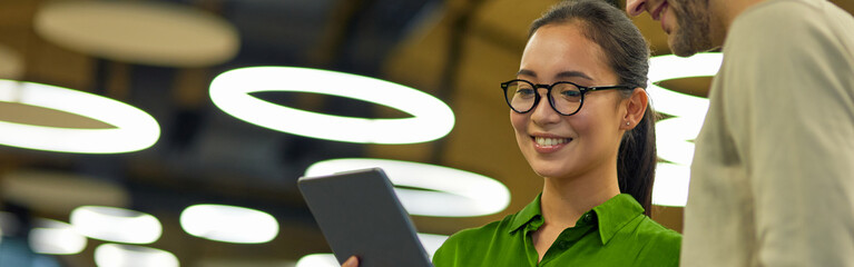 Young cheerful asian woman showing something on digital tablet to her male colleague while standing together in the coworking space or office