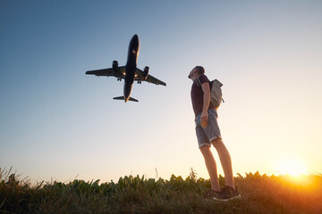 Man with backpack looking up to airplane landing at airport during beautiful summer sunset. .