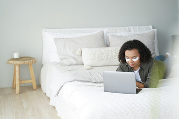 African-American teen with under-eye patches works on laptop in bed. Biracial lady shops online during morning skincare.
