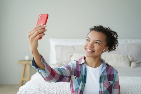 Biracial teen blogger chats online via phone video call, uses modern apps at home. Smiling girl takes selfie with smartphone.