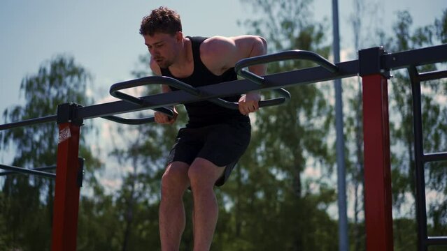 portrait of a young sportsman working out on parallel bars swinging his arms and shoulders doing push-ups on sports ground during street training