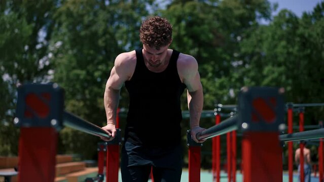 portrait of a young sportsman working out on parallel bars swinging his arms and shoulders doing push-ups on sports ground during street training