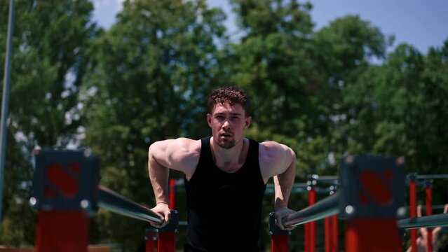 portrait of a young sportsman working out on parallel bars swinging his arms and shoulders doing push-ups on sports ground during street training