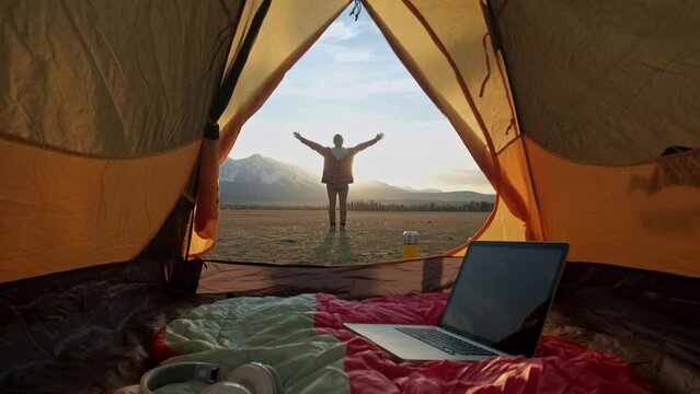 Man on mountain near orange tent looks into the distance hand up looking to the sunrise . Man in hood stands near an orange tent and looks at the bright sun. Summer landscape. The concept of privacy