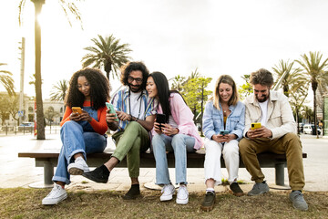 Multiethnic group of friends texting on their smartphones sitting on a bench in the street. Diverse young people smiling and using their phones outside in a park bench.