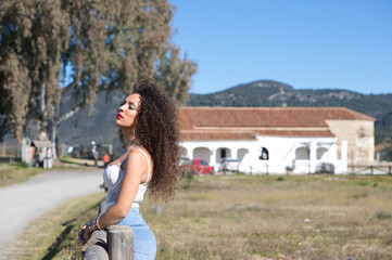 curly-haired, brunette, Spanish woman resting on the fence of the path leading to the meadow. She is dressed in jeans and a white top and enjoys the sunny day. The woman is sad and depressed.