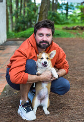 Young bearded man playing with his corgi dog in the forest in the morning