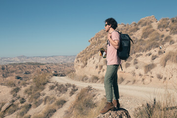 Cool young man in chino pants, pink tshirt and utility cargo backpack take little break from hiking on edge of road in deep dry canyon or valley. Hold hydration flask ready to drink water