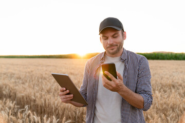 Bearded farmer with mobile phone and digital tablet in agricultural field at sunset..