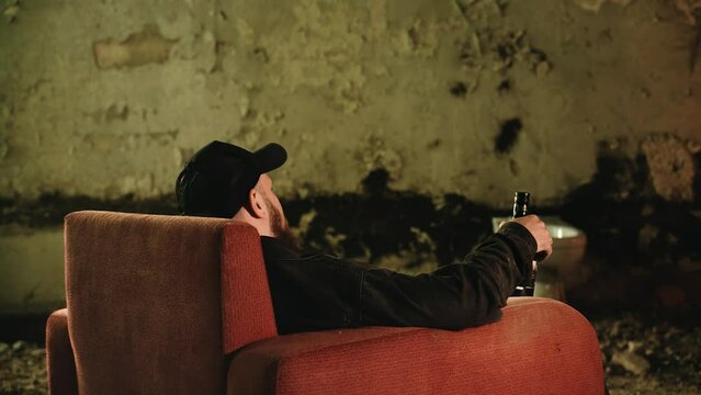 A man with a bottle sits down in a red armchair that stands in an abandoned house. Shooting in a brothel